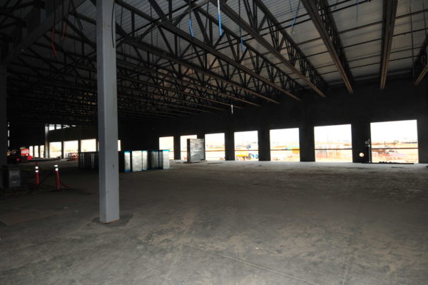 Large empty commercial building interior with concrete floor, exposed metal truss ceiling, and windows.