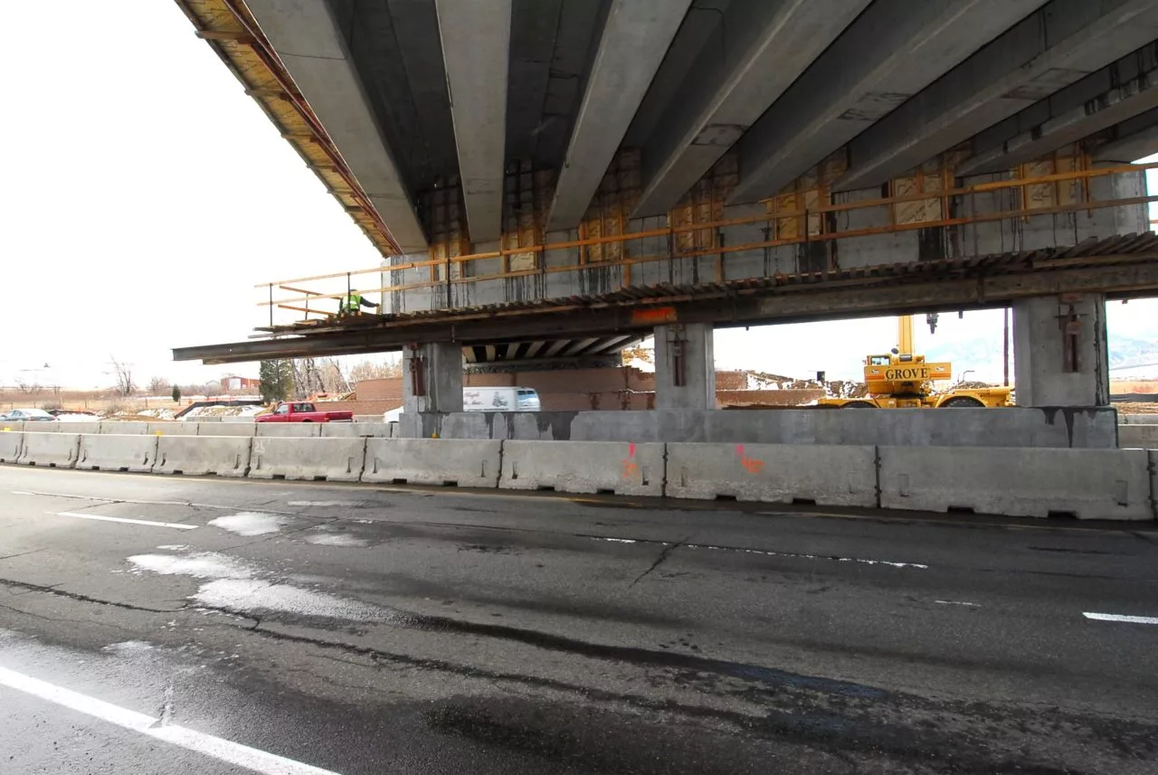 Highway under a concrete bridge undergoing construction, with barriers, a worker, and a crane visible.