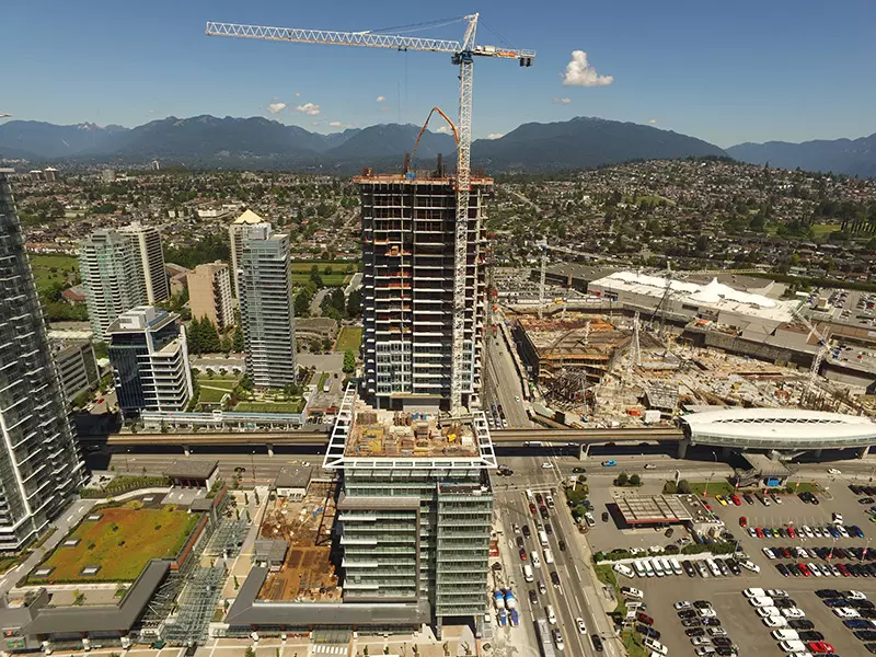 Aerial city view with a prominent skyscraper under construction, a crane, sprawling urban development, and distant mountains.