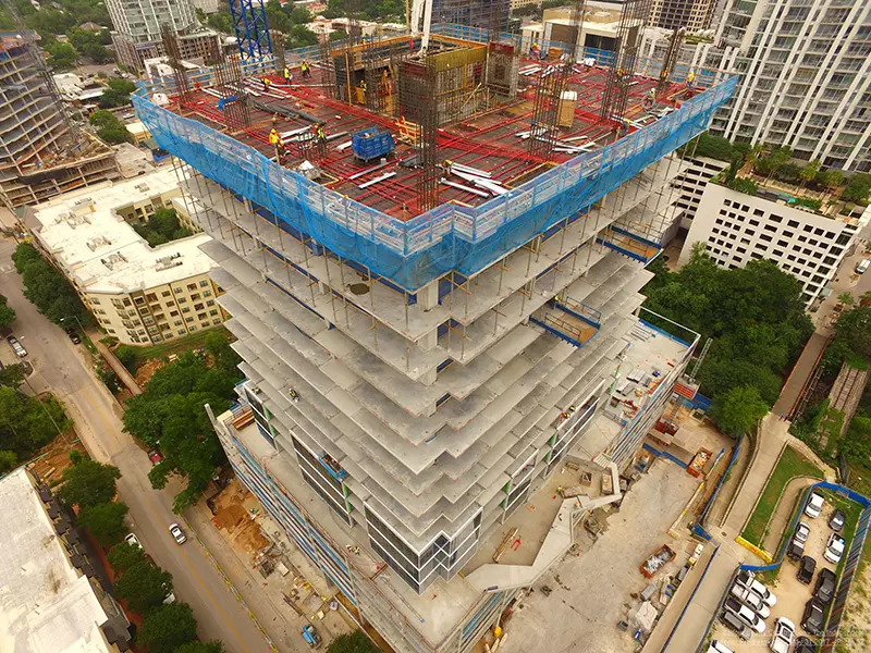 Aerial view of a high-rise building under construction, showing concrete floors, rebar, workers, and safety netting.