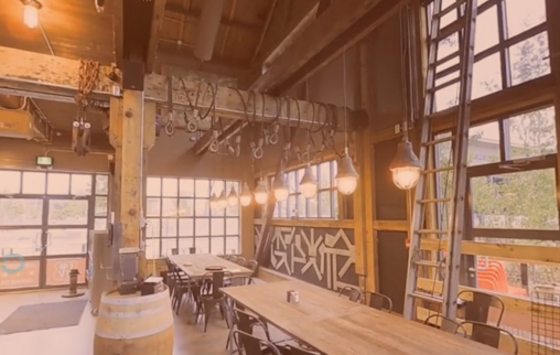 Industrial-style restaurant interior with long wooden tables, pendant lights, exposed beams, and large grid windows.