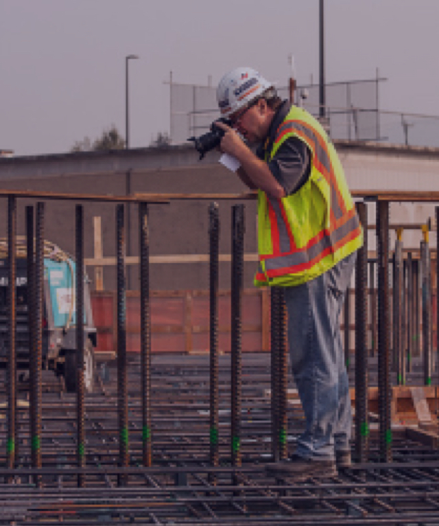 Construction worker in hard hat and safety vest photographing rebar on a job site.