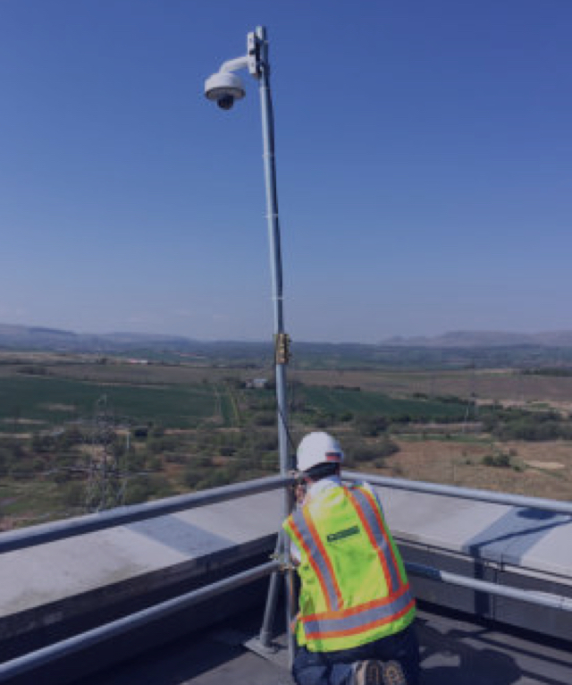 Worker in hard hat installing a security camera system on a pole from a rooftop, overlooking a rural landscape.