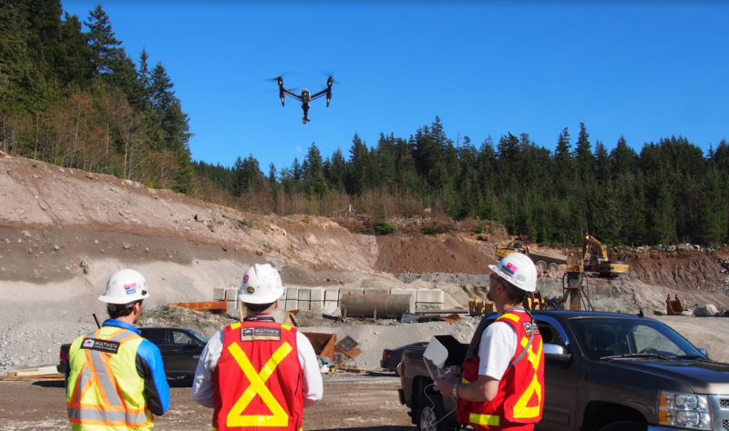 Three construction workers in hard hats monitor a drone flying over a job site.