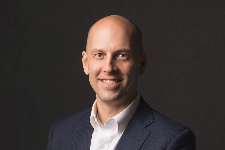 Professional headshot of a smiling bald man in a dark pinstripe suit and white shirt.