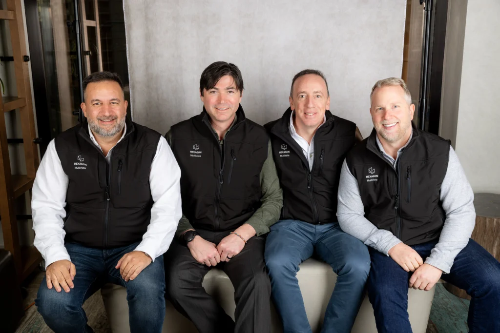 Four smiling men in HEXAGON Multivista vests seated for a team photo.