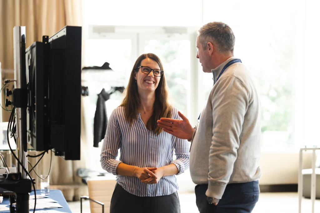 Smiling woman in glasses listens intently to a gesturing man at a professional event or office discussion.