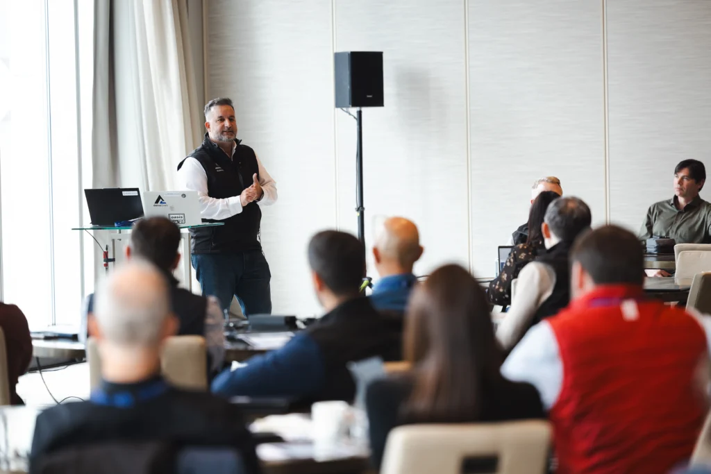 A speaker presents to an audience at a business conference, gesturing with his hands.