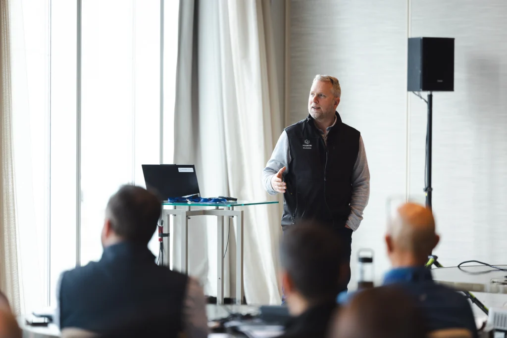A man in a HEXAGON Multivista vest speaks to an audience during a presentation.