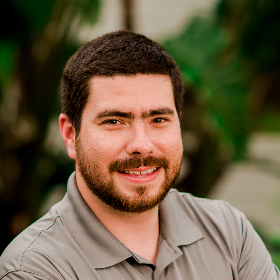Headshot of a smiling man with a beard and short dark hair wearing a gray polo shirt, blurred green background.