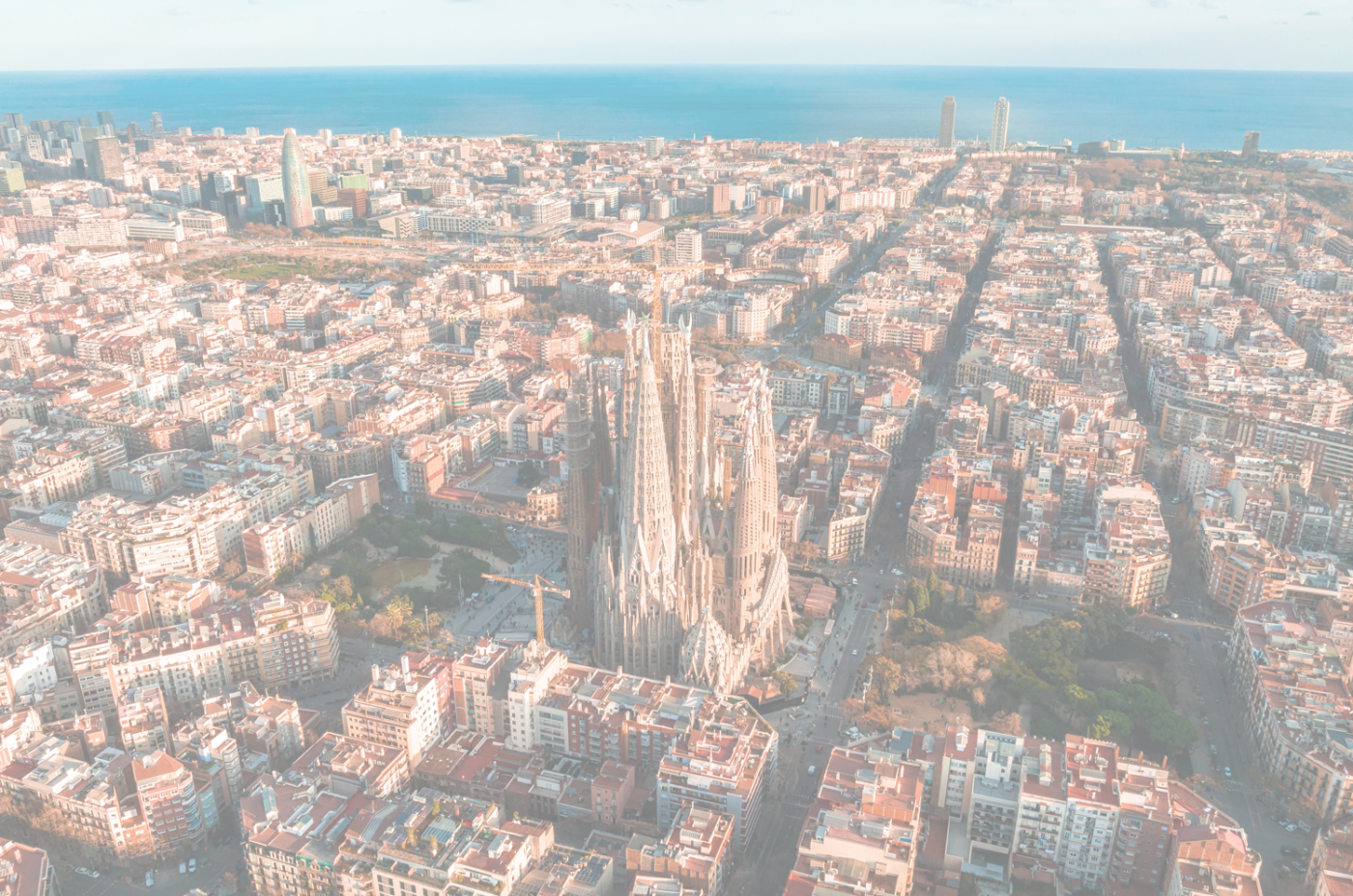 Aerial view of Barcelona, showcasing the iconic Sagrada Familia cathedral surrounded by dense city blocks and the sea.