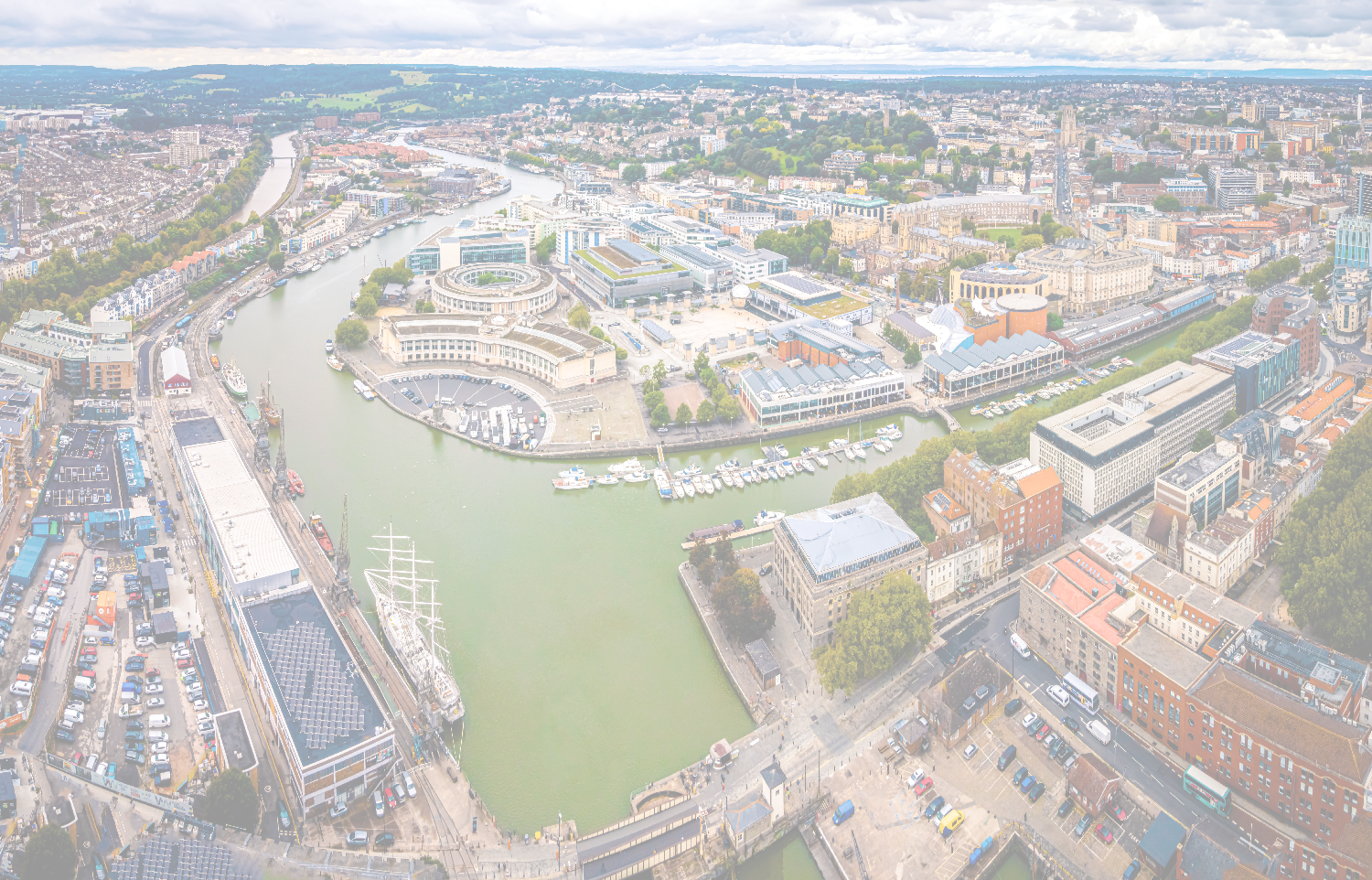 Aerial view of a city's winding harbor with historic buildings, modern structures, and multiple ships, including a tall ship.