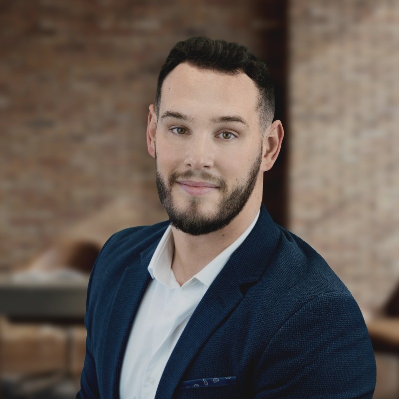 Headshot of a smiling bearded man in a blue blazer against a blurred brick background.