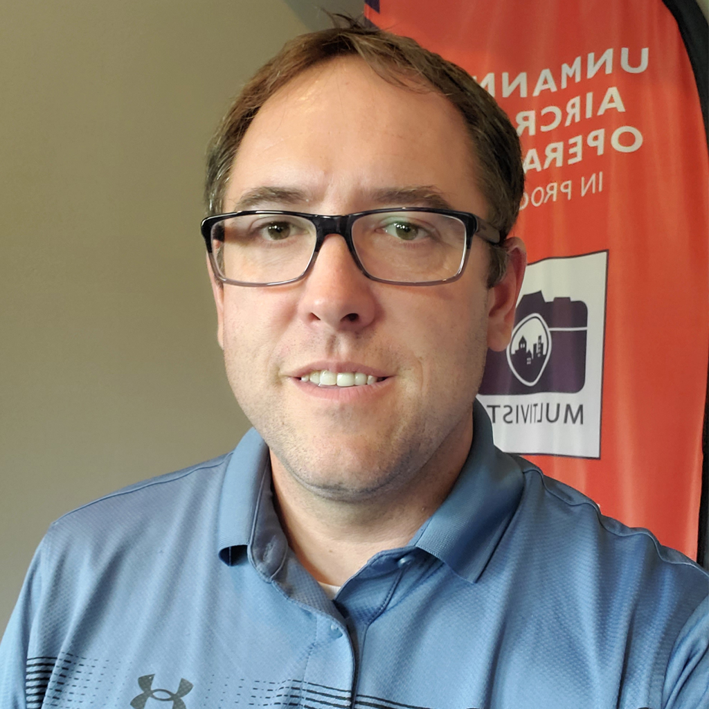 Man with glasses smiling in a blue polo shirt, with an orange banner behind him.