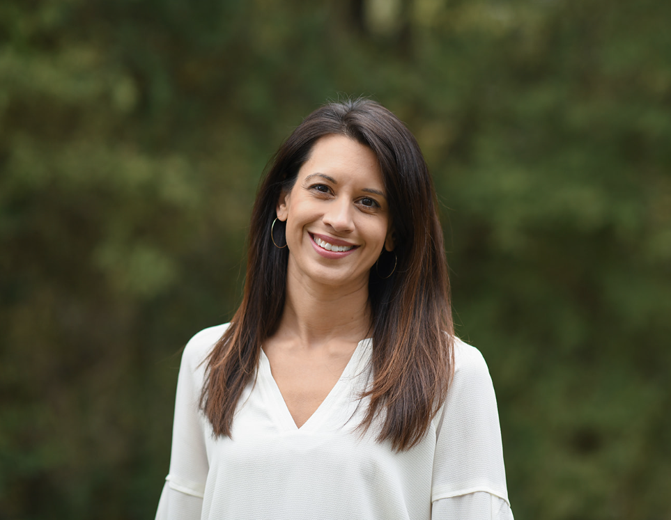 Smiling woman with long dark hair and hoop earrings in a white blouse, blurry green background.