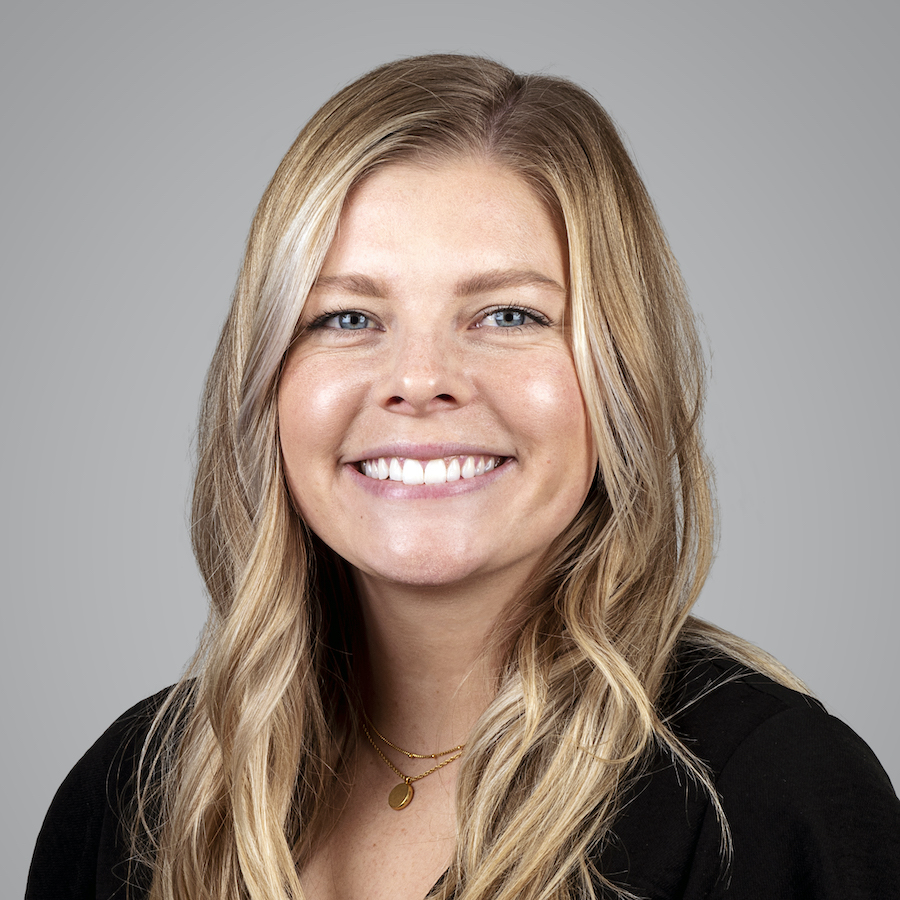 Smiling young woman with blonde hair, blue eyes, and a gold necklace against a gray background.