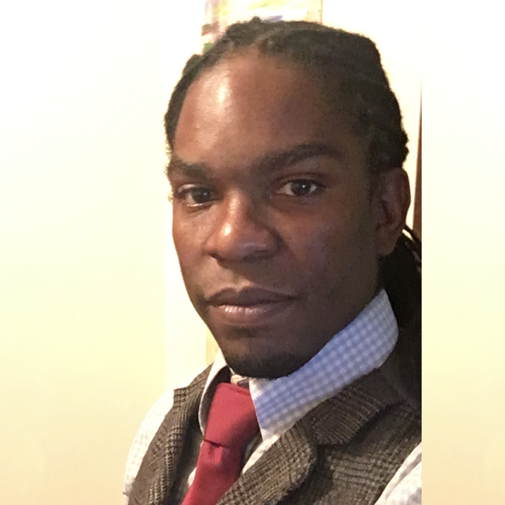 Professional headshot of a Black man with dreadlocks, wearing a plaid vest and red tie.