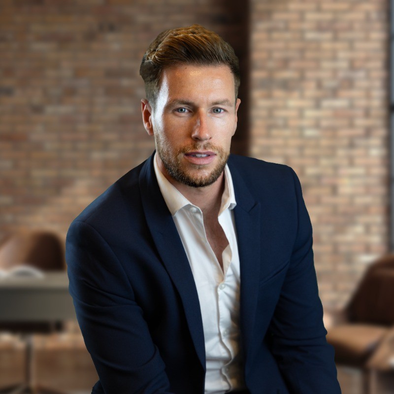 Professional portrait of a bearded man in a navy suit and white shirt against a blurred brick wall background.