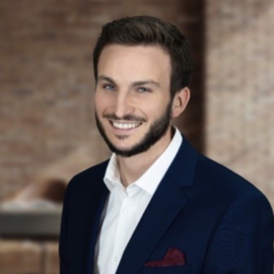 Smiling man with blue eyes, dark hair, and beard, wearing a navy suit with a maroon pocket square.