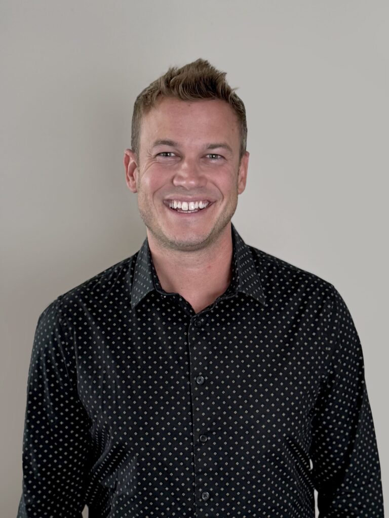 Headshot of a smiling man with short brown hair wearing a black patterned shirt.