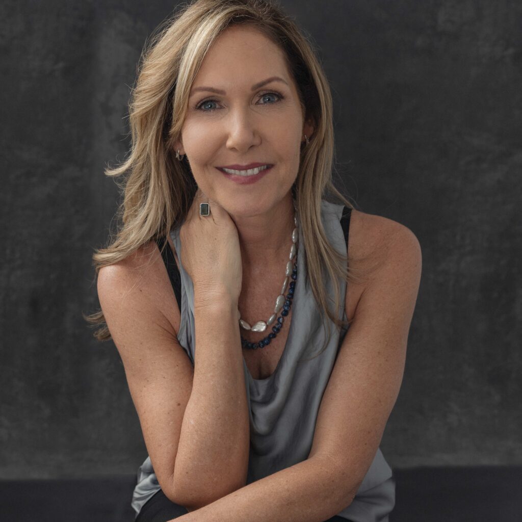 Smiling woman with blonde hair, grey top, and pearl necklace, hand on chin, dark background.