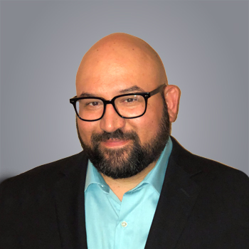 Smiling man with beard, glasses, blue shirt, and black jacket.