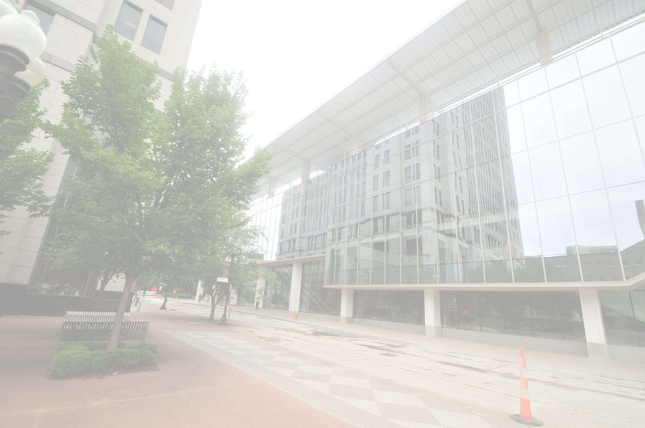 Modern glass building on a city street with trees, benches, and streetlights.