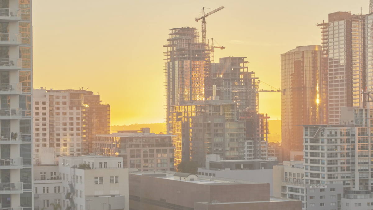 Golden sunset bathes a city skyline featuring modern buildings and towering construction cranes.