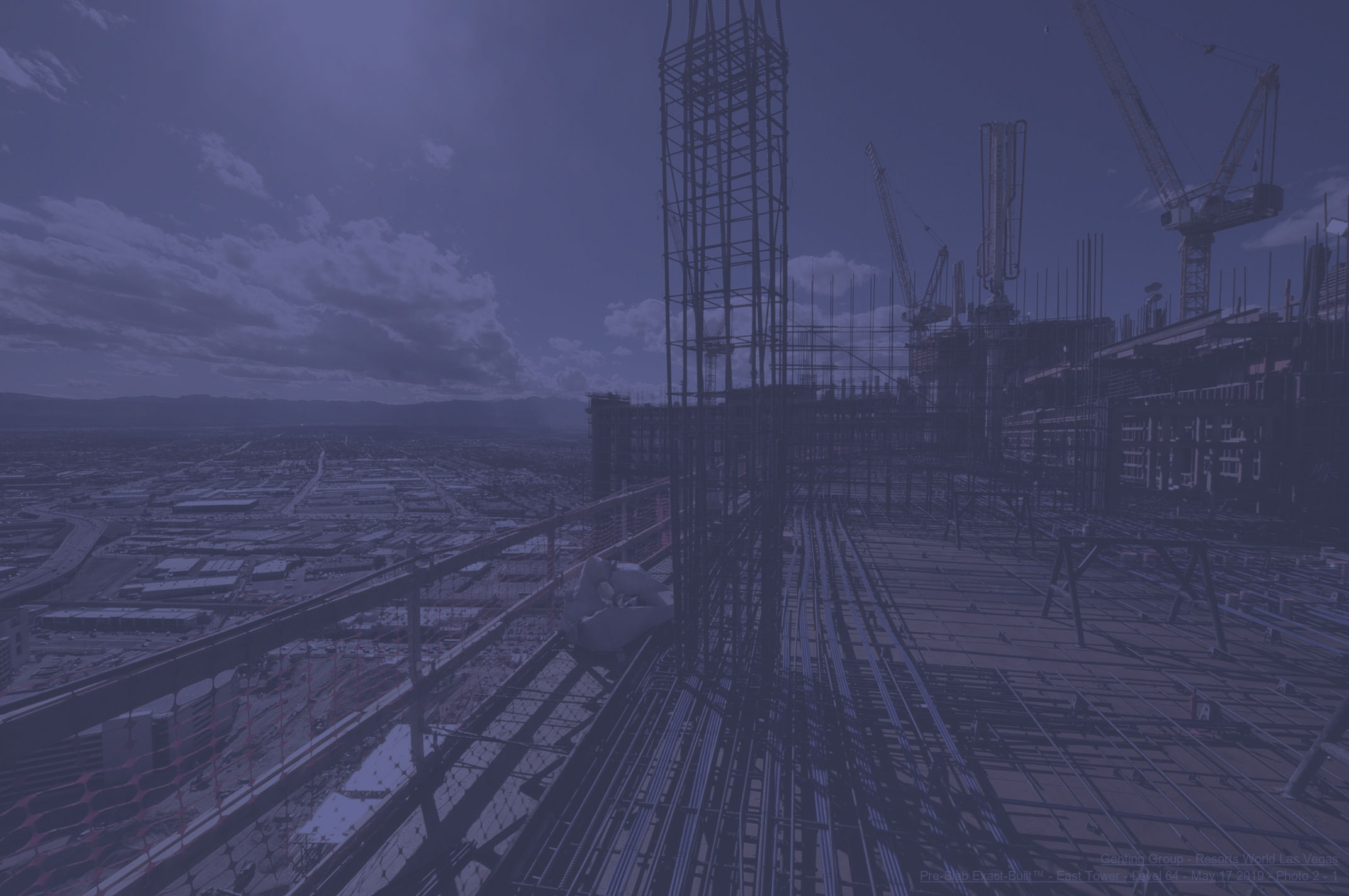 High-angle view of skyscraper construction: rebar on upper floor, cranes, and a vast city stretching to distant mountains.
