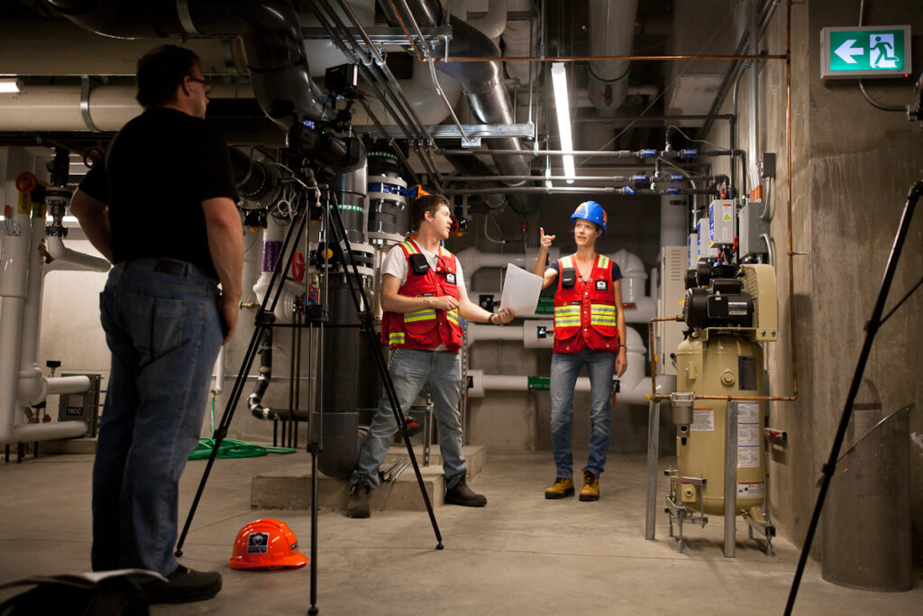 Two workers in safety vests film a video, discussing machinery in a mechanical room.