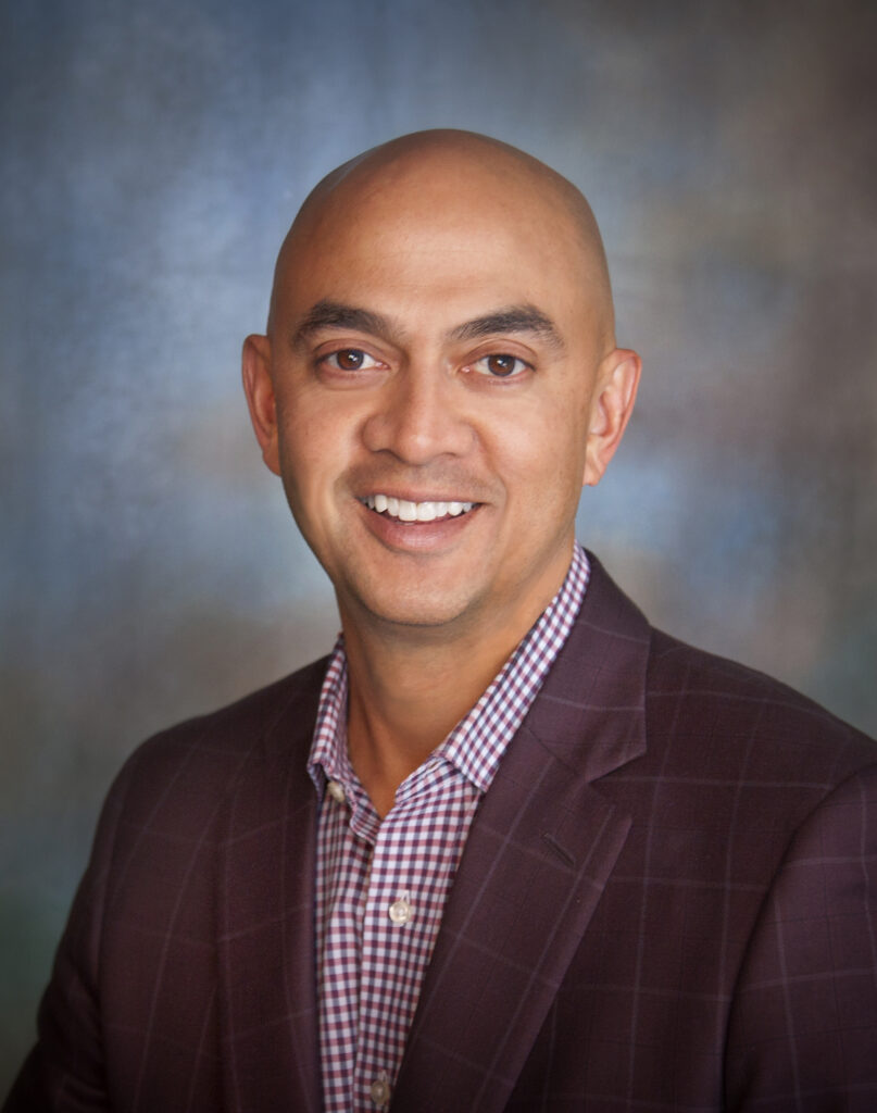 Headshot of a smiling bald man in a burgundy plaid suit jacket and checkered shirt.