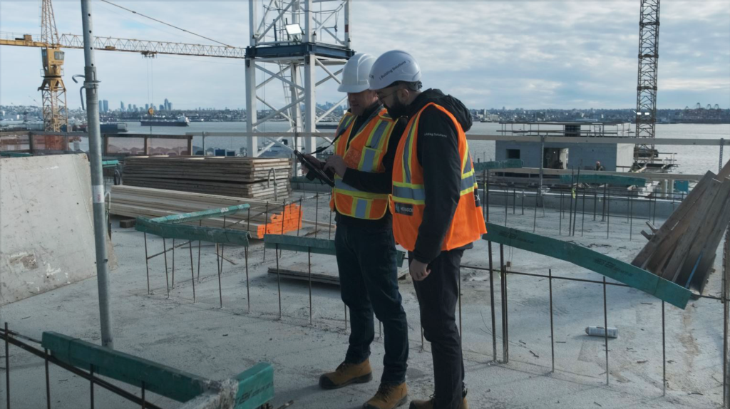 Two engineers in hard hats and safety vests review a tablet on a construction site overlooking a city.