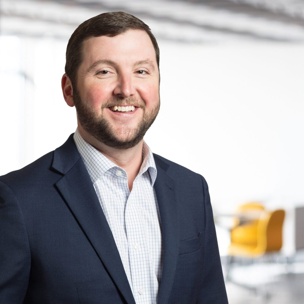 Smiling bearded man in a blue suit jacket and checkered shirt, professional headshot.