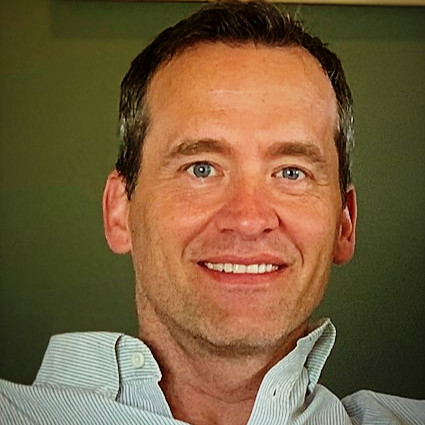 Smiling man with blue eyes and dark hair wearing a light striped shirt, against a green background.
