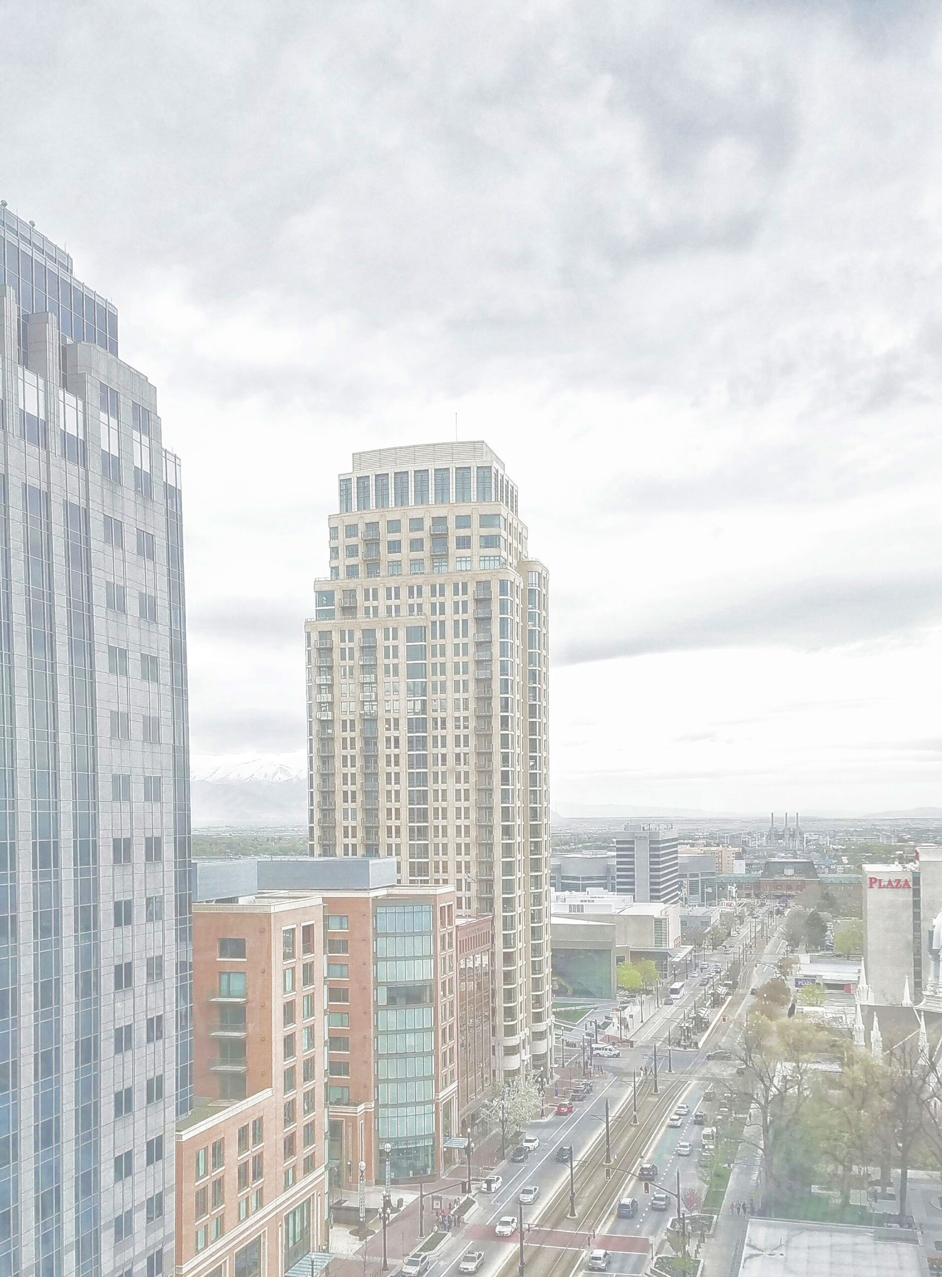 Aerial view of a city with modern skyscrapers, a busy street, and distant mountains under a gray sky.