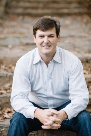 Man smiling in a light blue shirt and jeans, sitting on outdoor stone steps.
