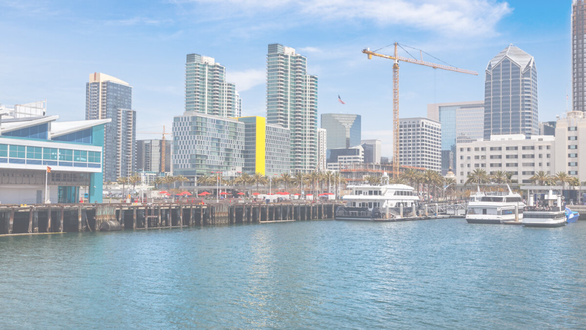 Harbor view of a city skyline with modern high-rises, docked boats, palm trees, and a construction crane.