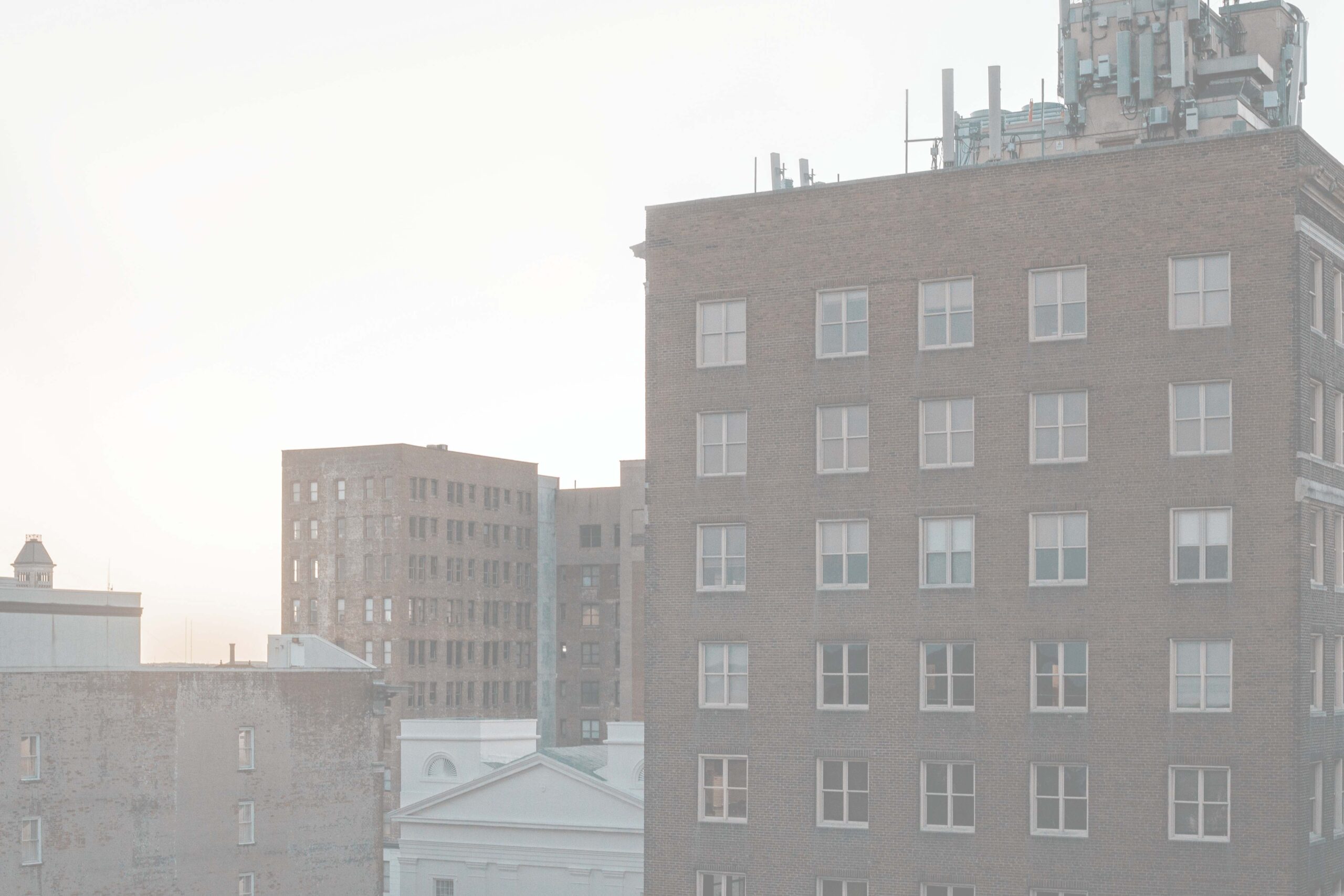Hazy urban skyline with brick buildings, many windows, and rooftop cellular antennas against a bright, washed-out sky.
