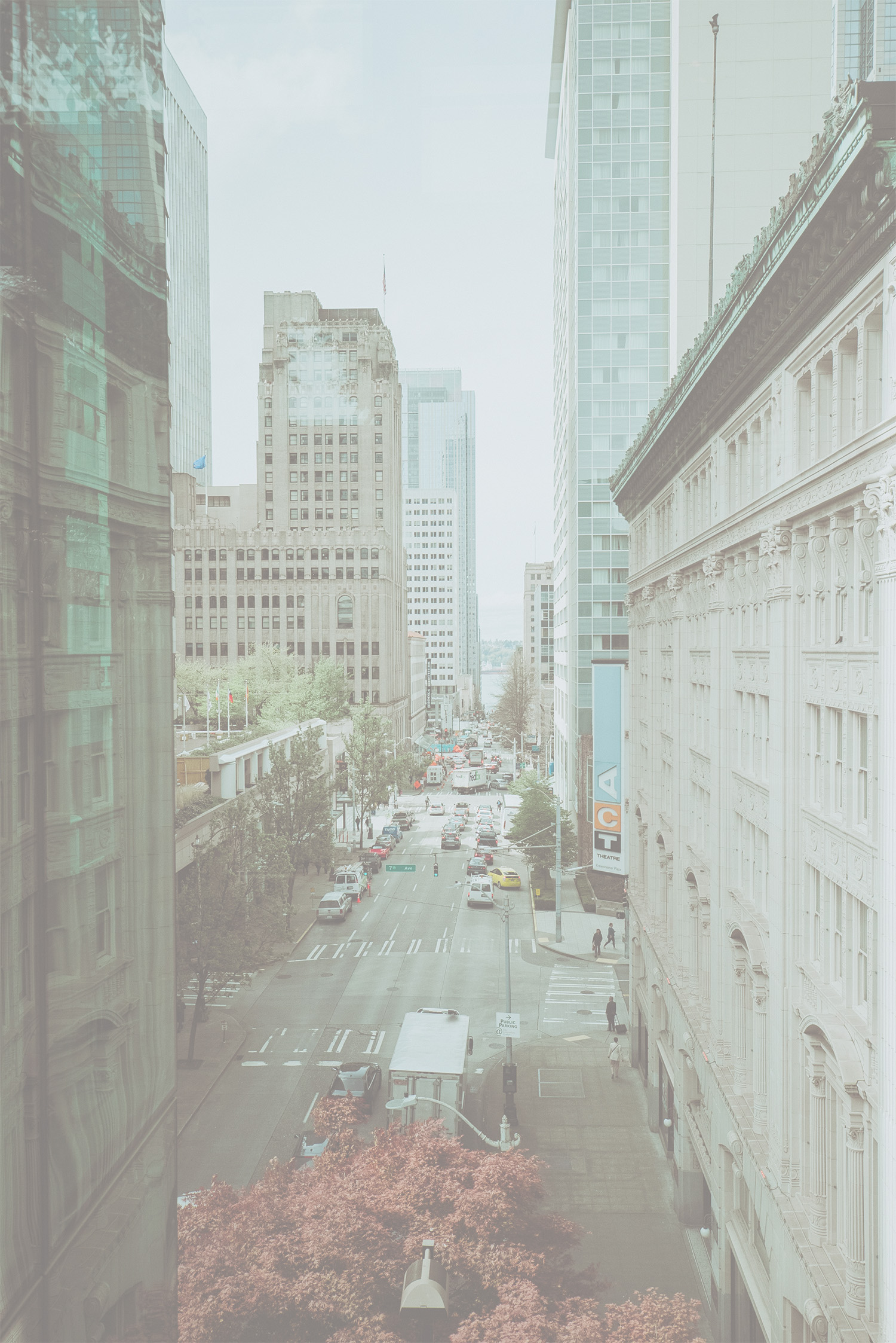 Busy city street lined with tall buildings, cars, and pedestrians, with a glimpse of water in the distance.