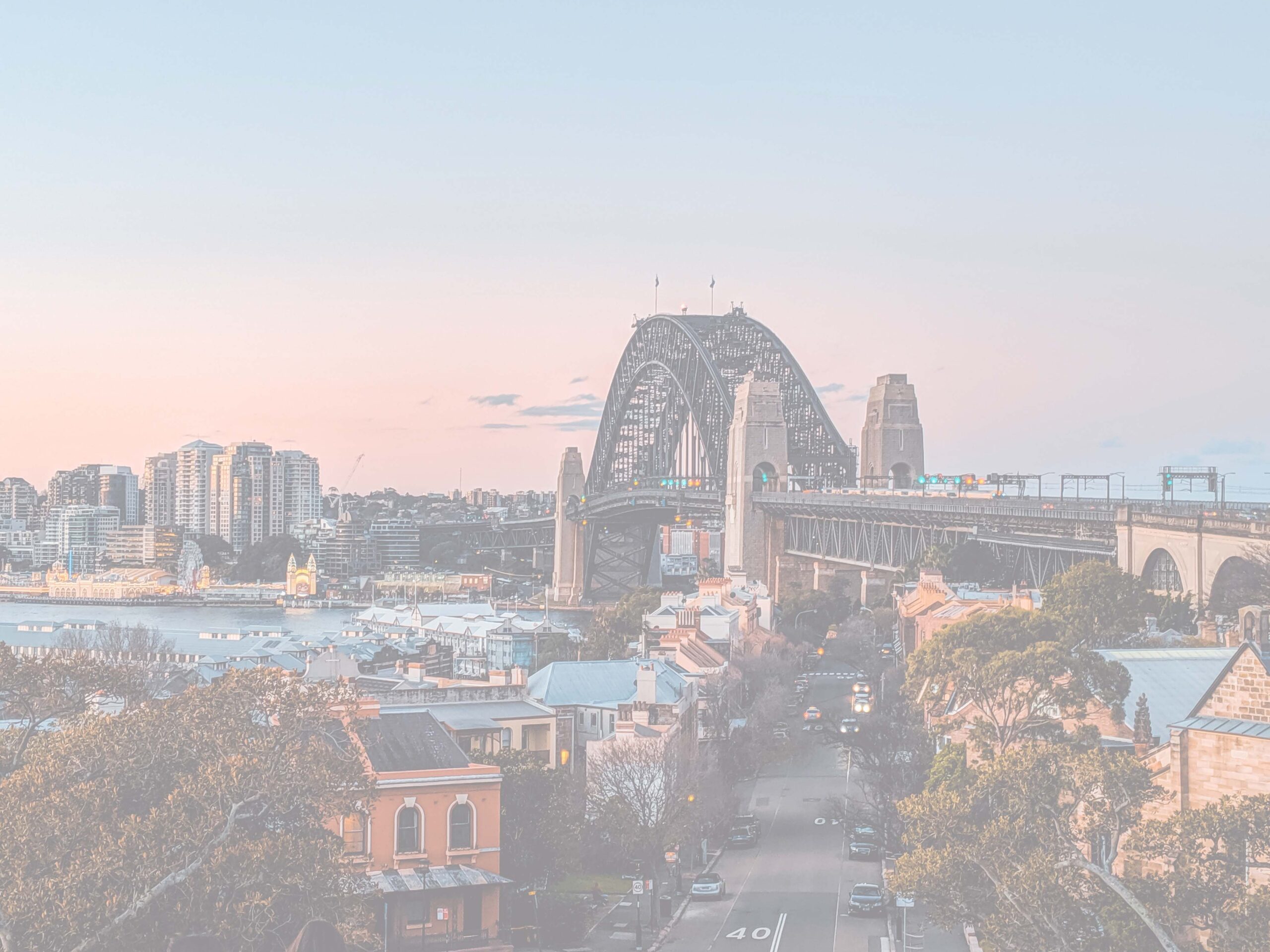 Aerial view of Sydney Harbour Bridge and city at pastel dusk, with streets and harbor below.