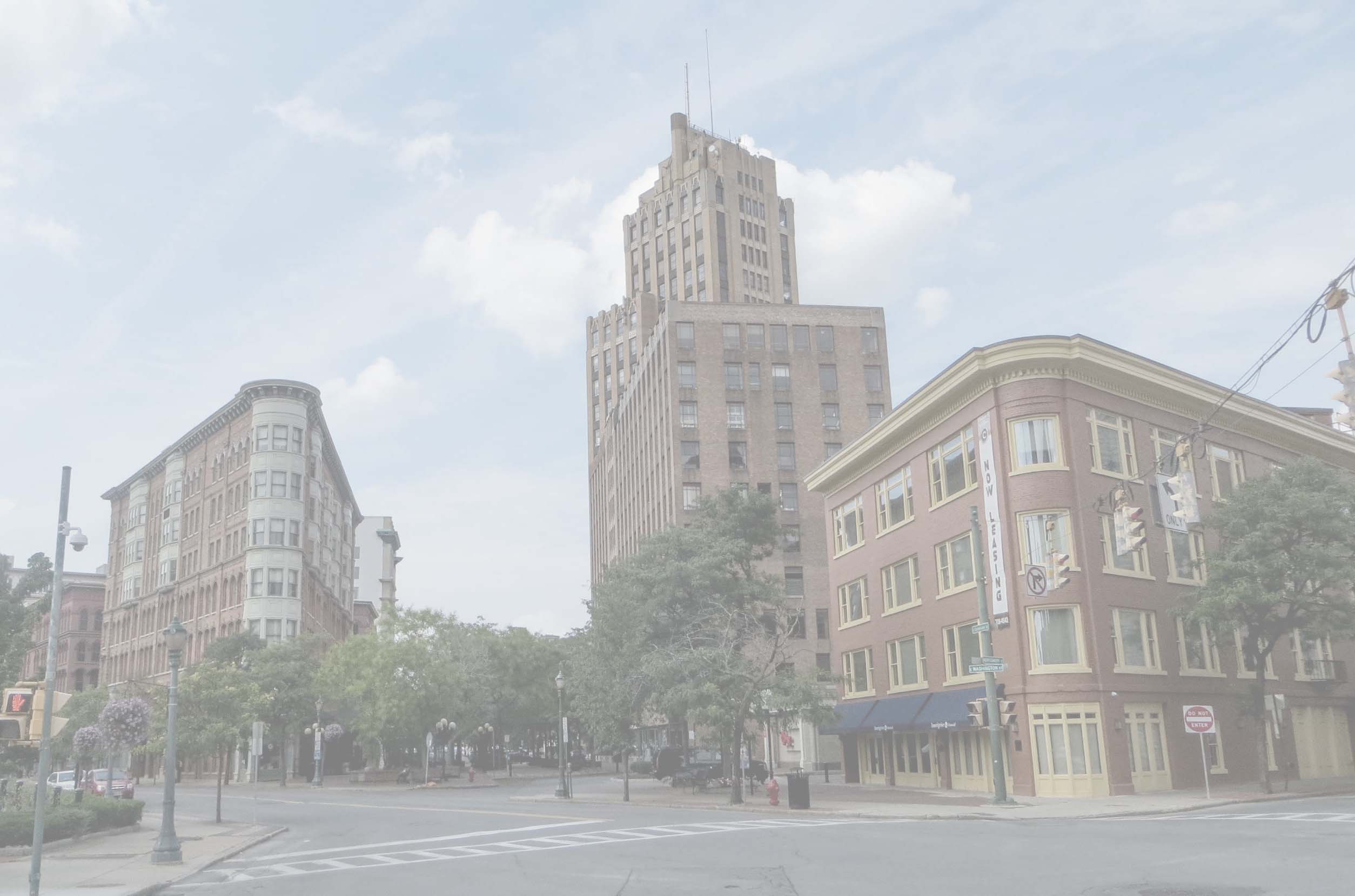 Historic city corner with brick buildings, a tall skyscraper, street, traffic lights, and green trees.