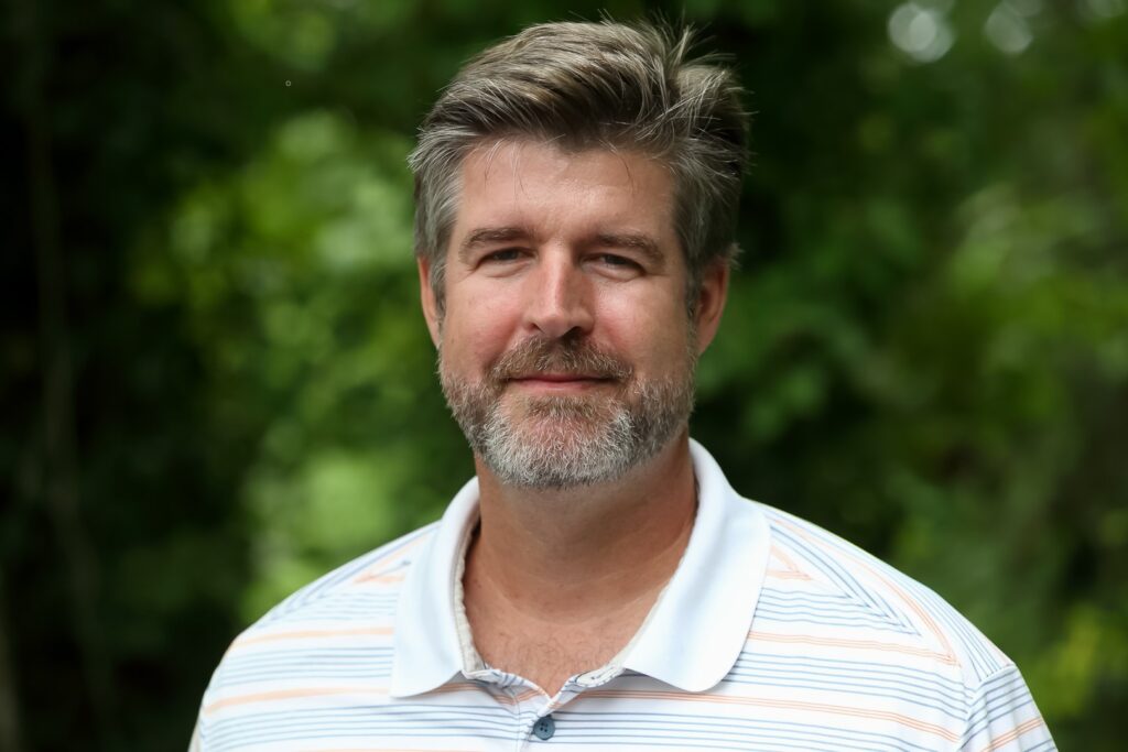 Headshot of a smiling middle-aged man with grey hair and beard, wearing a striped polo shirt outdoors.