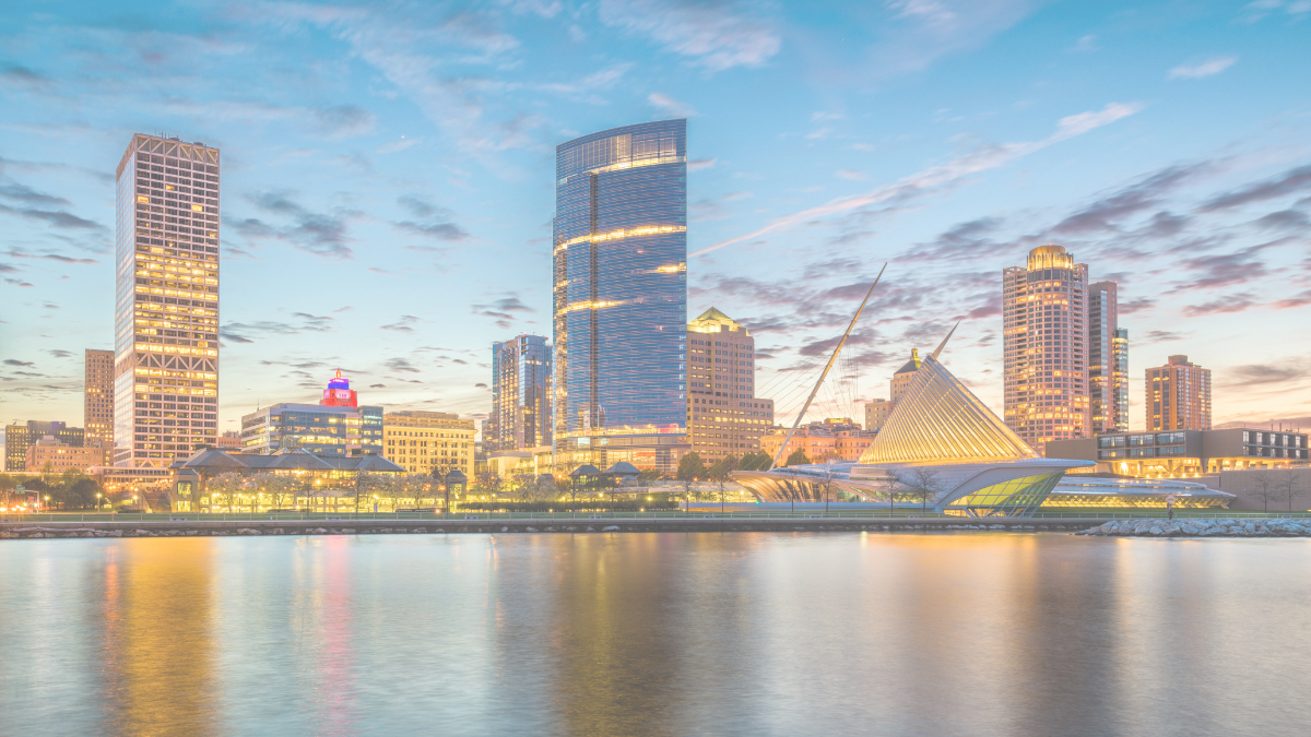 Milwaukee skyline at sunset, showing illuminated skyscrapers, the Art Museum, and reflections on Lake Michigan.