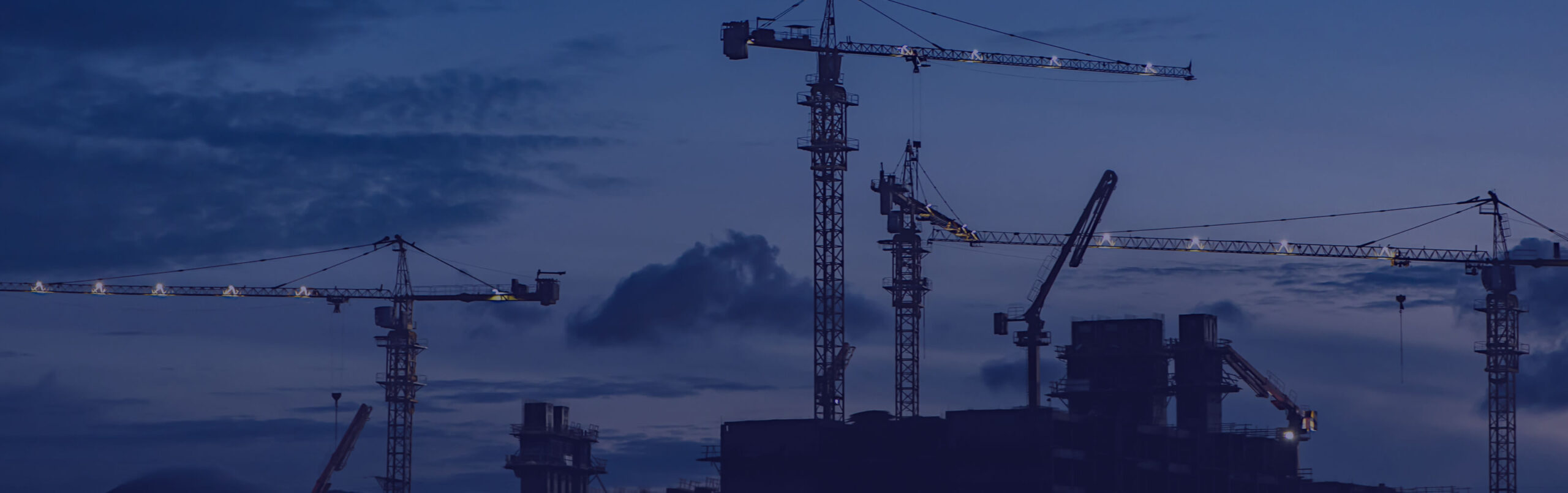 Construction site with multiple cranes silhouetted against a cloudy twilight sky.