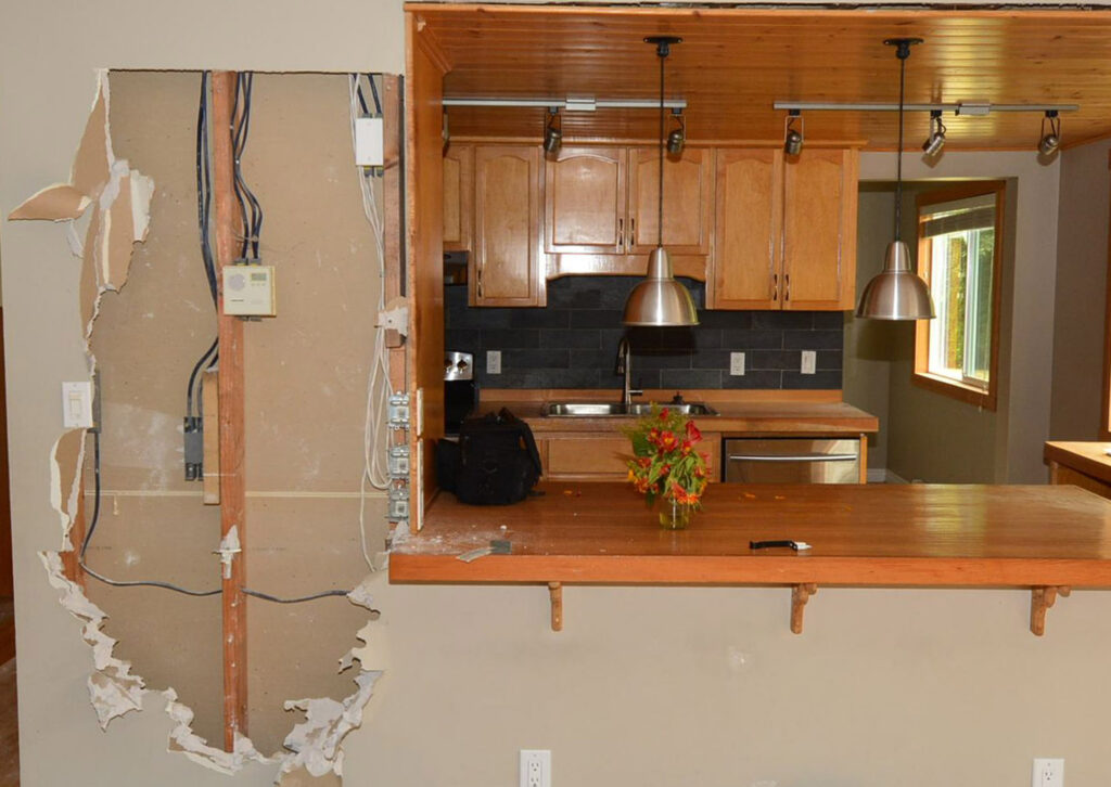 Kitchen renovation with drywall removed exposing wiring on the left, contrasting with a functional kitchen counter and cabinets on the right.