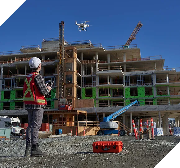Engineer in safety vest controls a drone over a multi-story building under construction.
